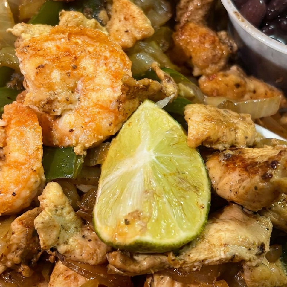 Grilled chicken and shrimp with sautéed peppers and onions, lime wedge, and black beans on a plate.